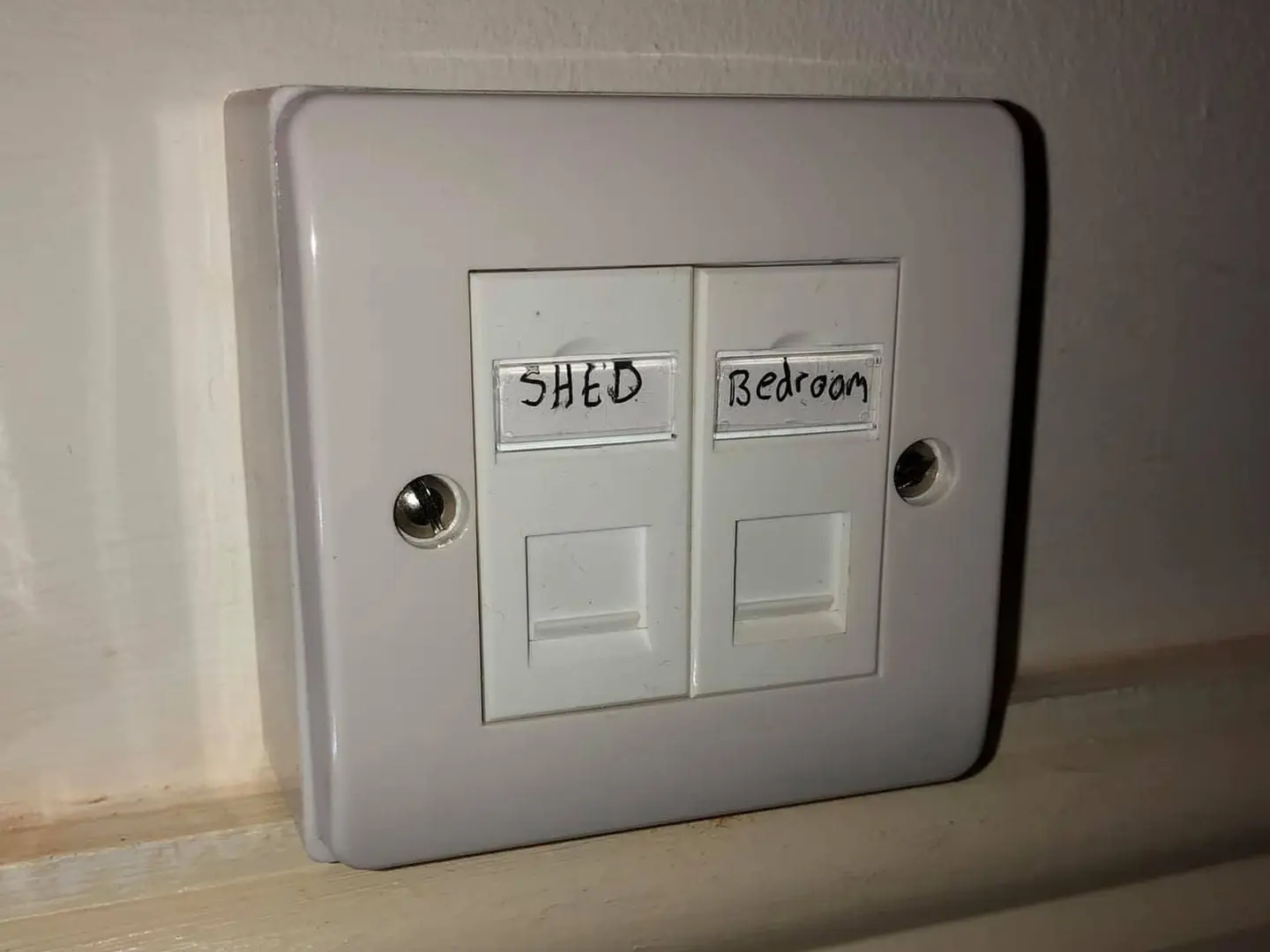 Twin ethernet faceplate hand-labelled SHED and Bedroom on a white surface box resting on a skirting board