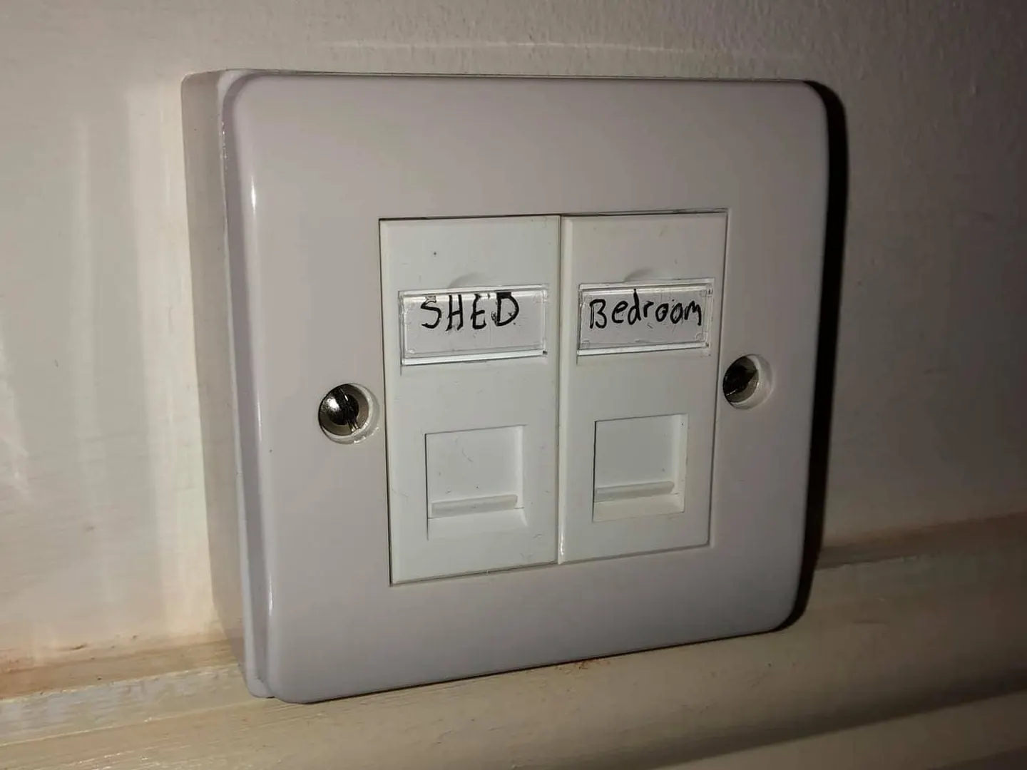 Twin ethernet faceplate hand-labelled SHED and Bedroom on a white surface box resting on a skirting board