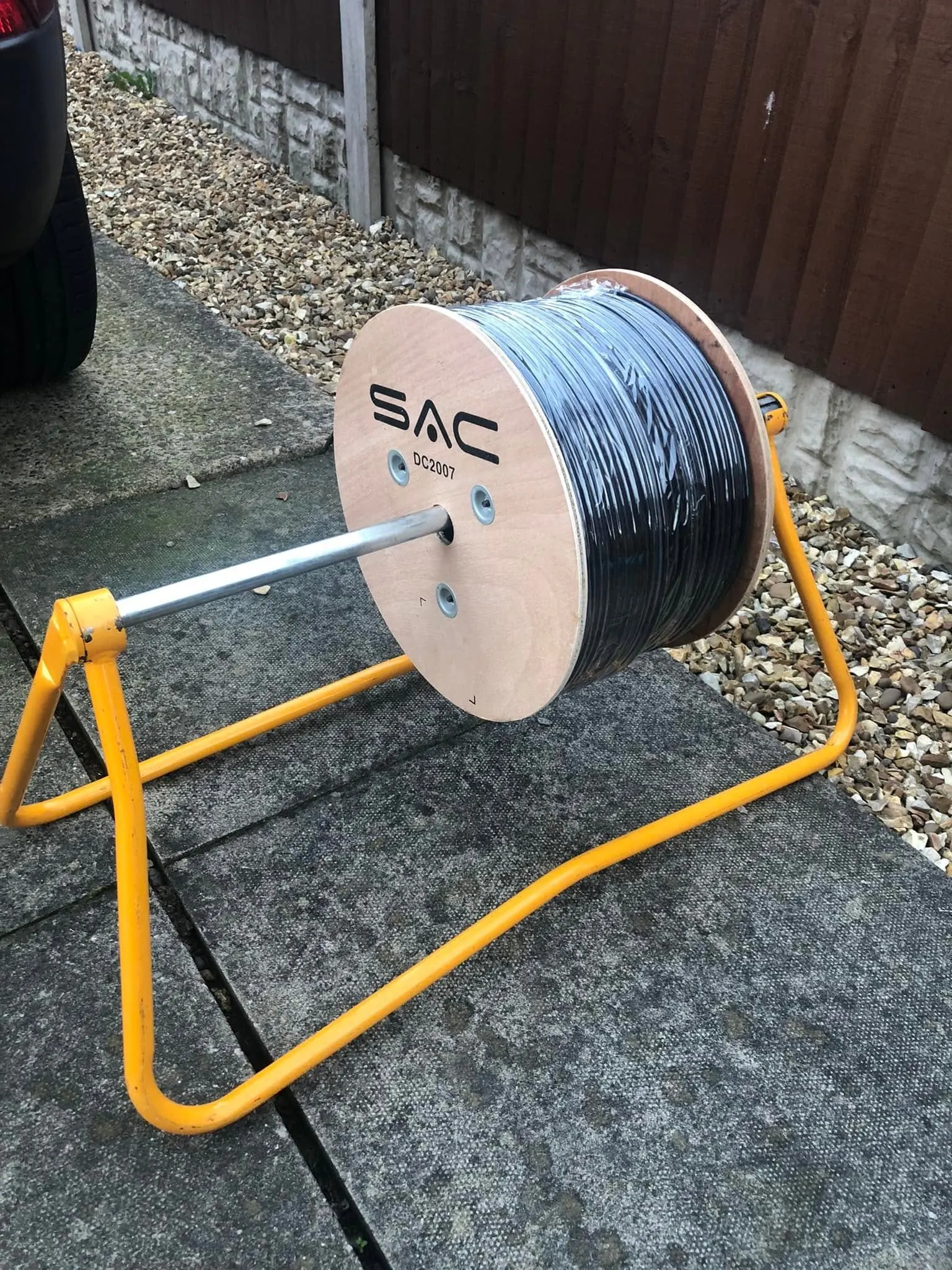 Drum of black cable still in its wrap on a yellow reel stand, set on paving slabs beside a gravel border and fence