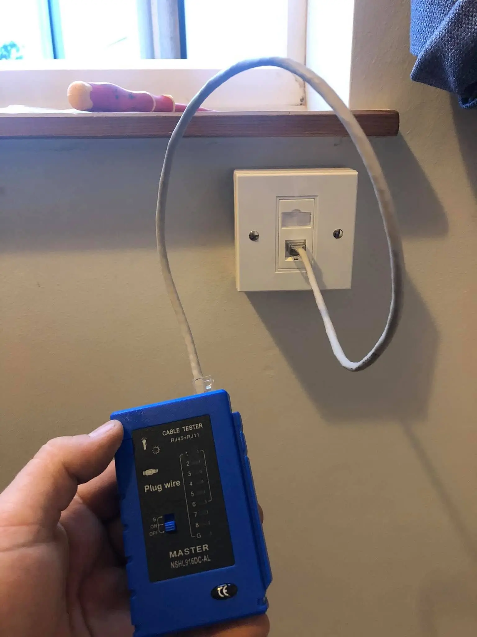 Hand holding a blue RJ45 cable tester connected by a grey patch lead to a white data faceplate under a windowsill