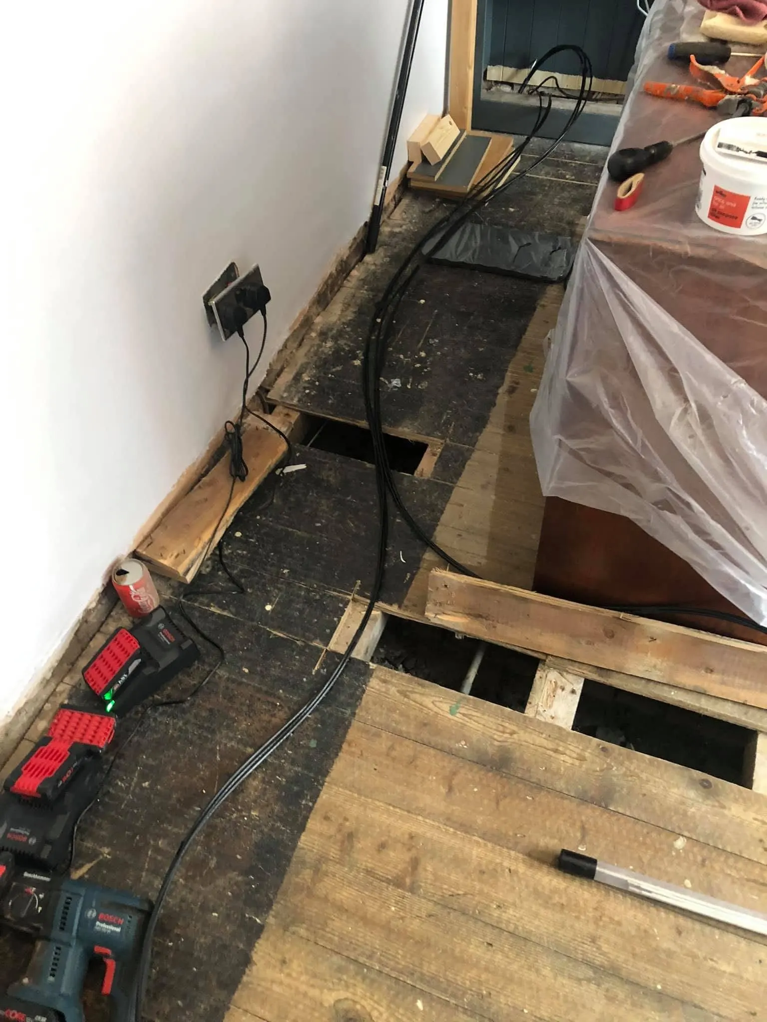 Black cables laid along a floor with boards lifted over the joists, next to Bosch batteries and a cordless drill