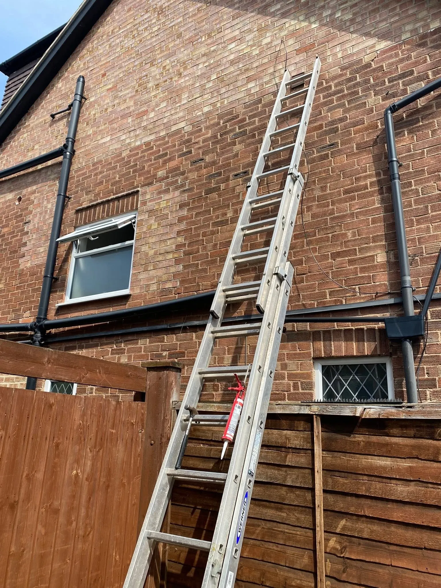 Aluminium extension ladder against a brick gable wall with a claw hammer and silicone gun hung on its rungs and a black cable up the wall