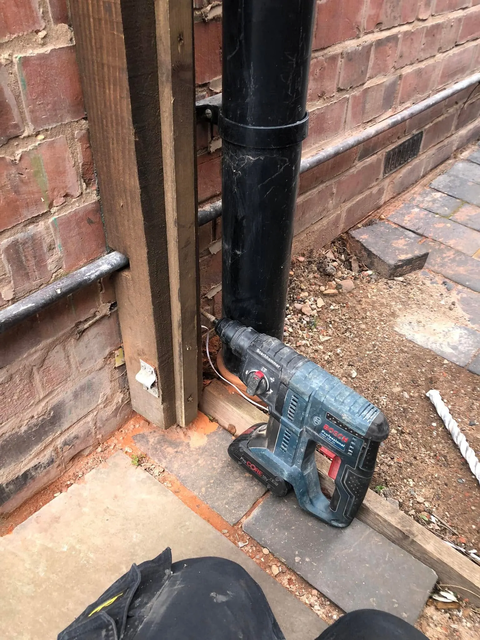 Bosch cordless SDS drill resting against a black downpipe at the base of a red brick wall, with a thin white cable beside it
