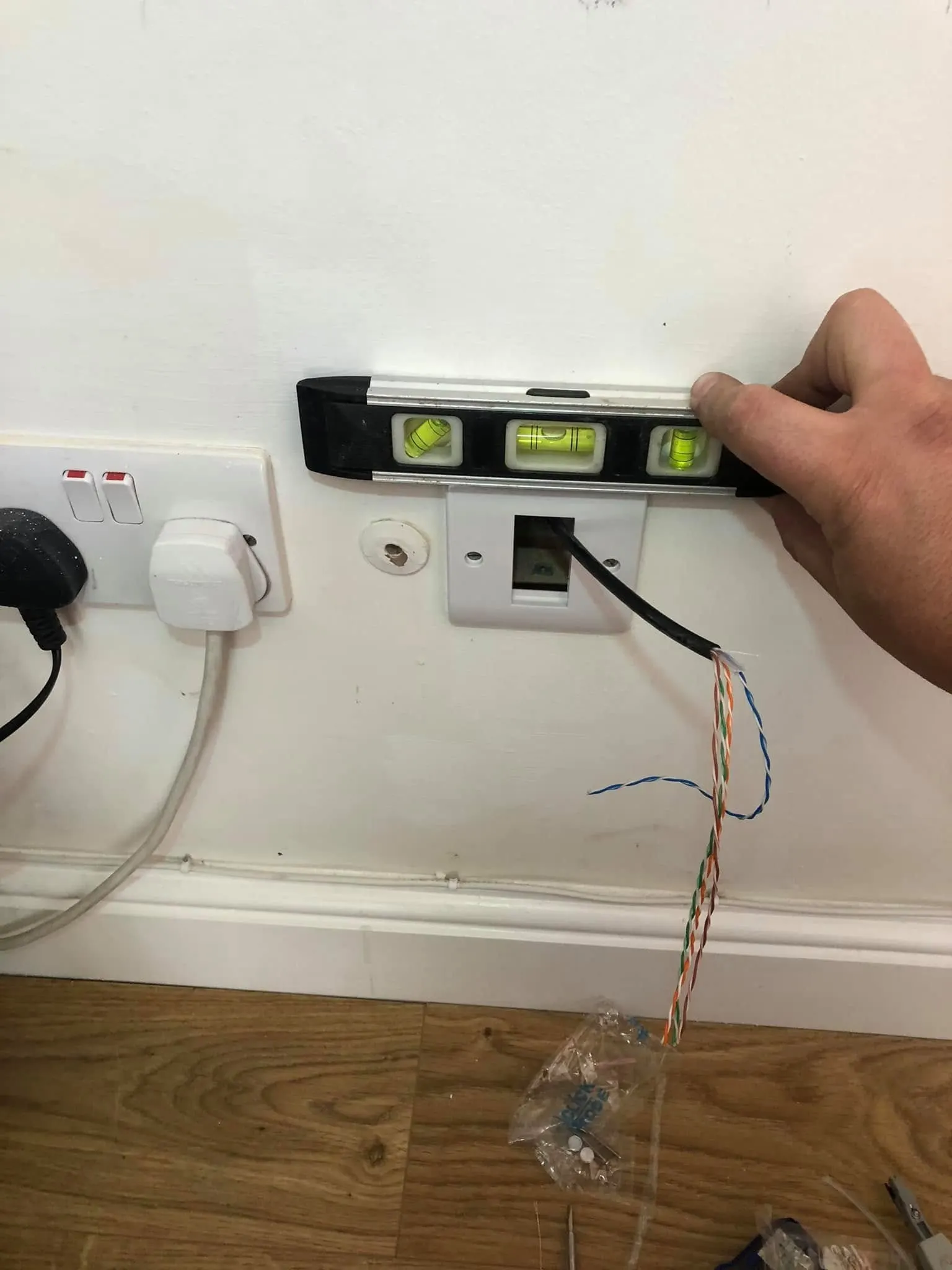 Hand holding a torpedo spirit level on top of a white data faceplate with a stripped black cable hanging below