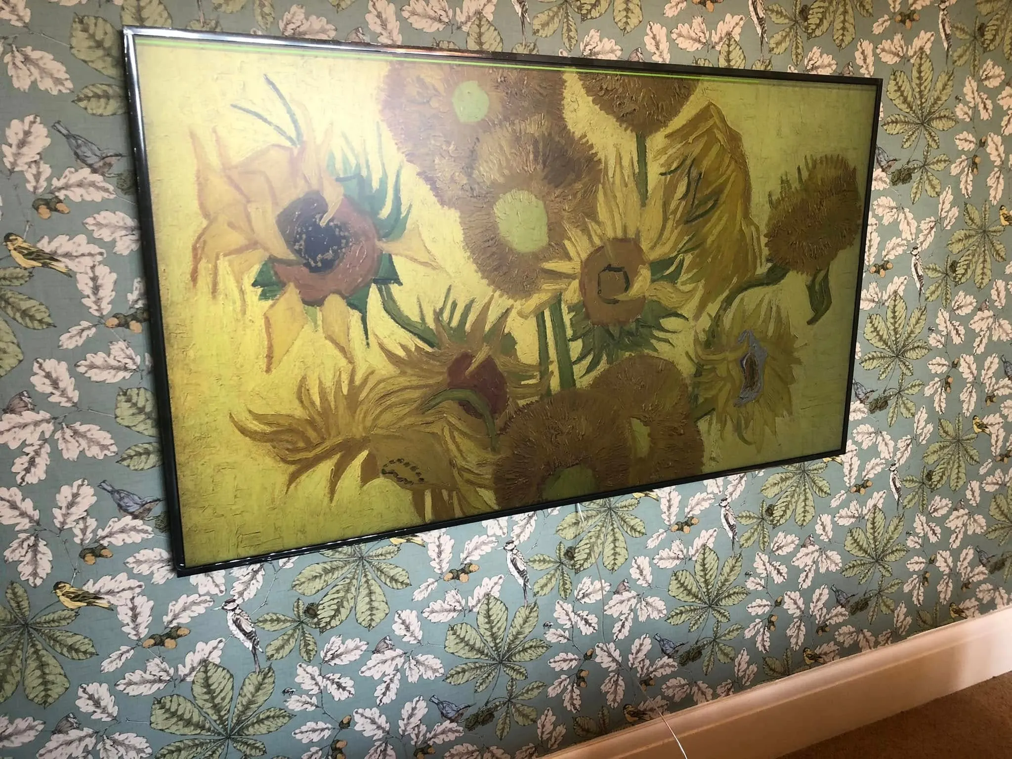 TV showing a sunflower painting hangs close to botanical wallpaper, seen from an angle above the skirting board