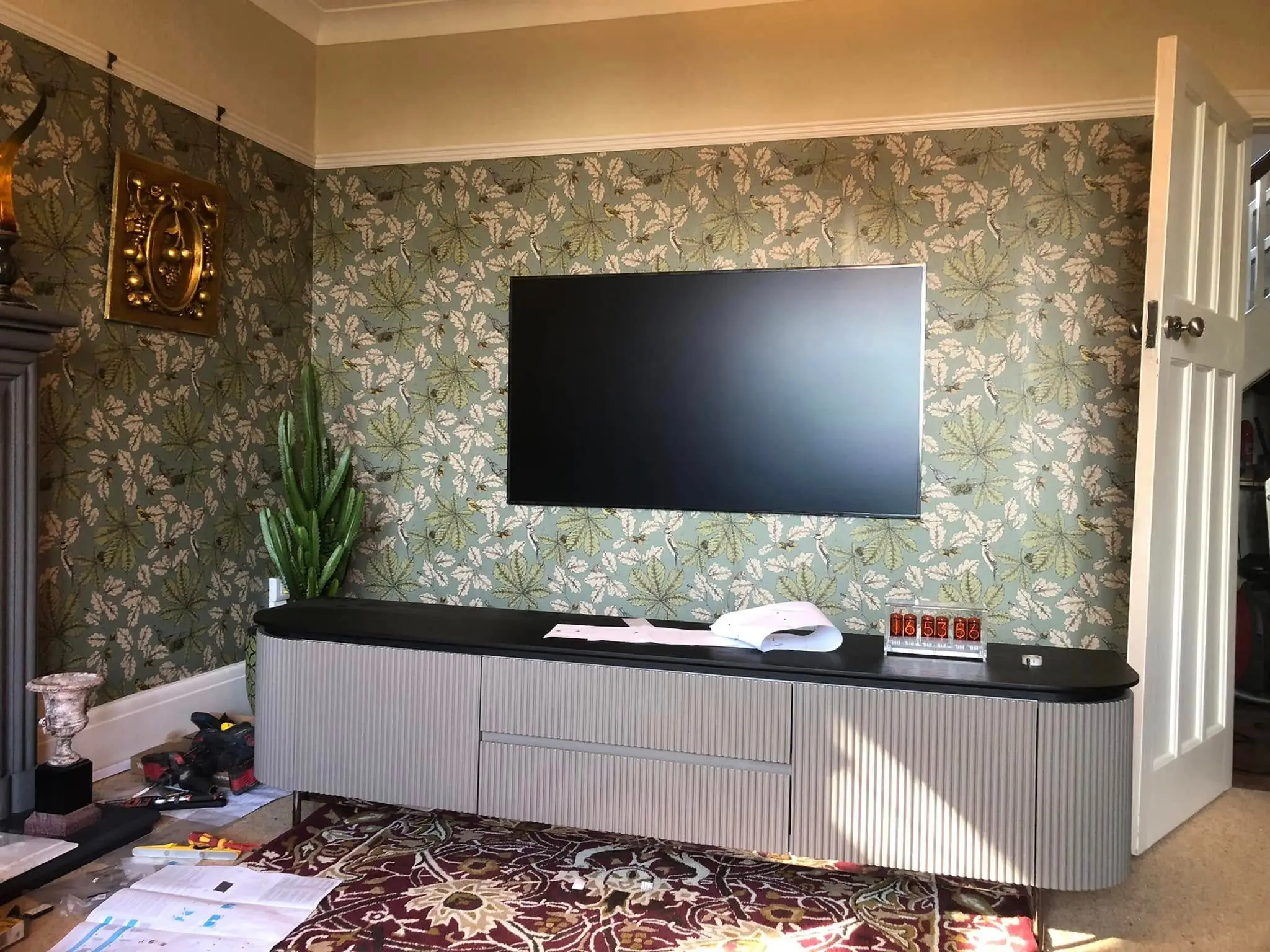 Wall-mounted TV sitting flat against green botanical wallpaper above a long grey fluted sideboard, with tools on the floor