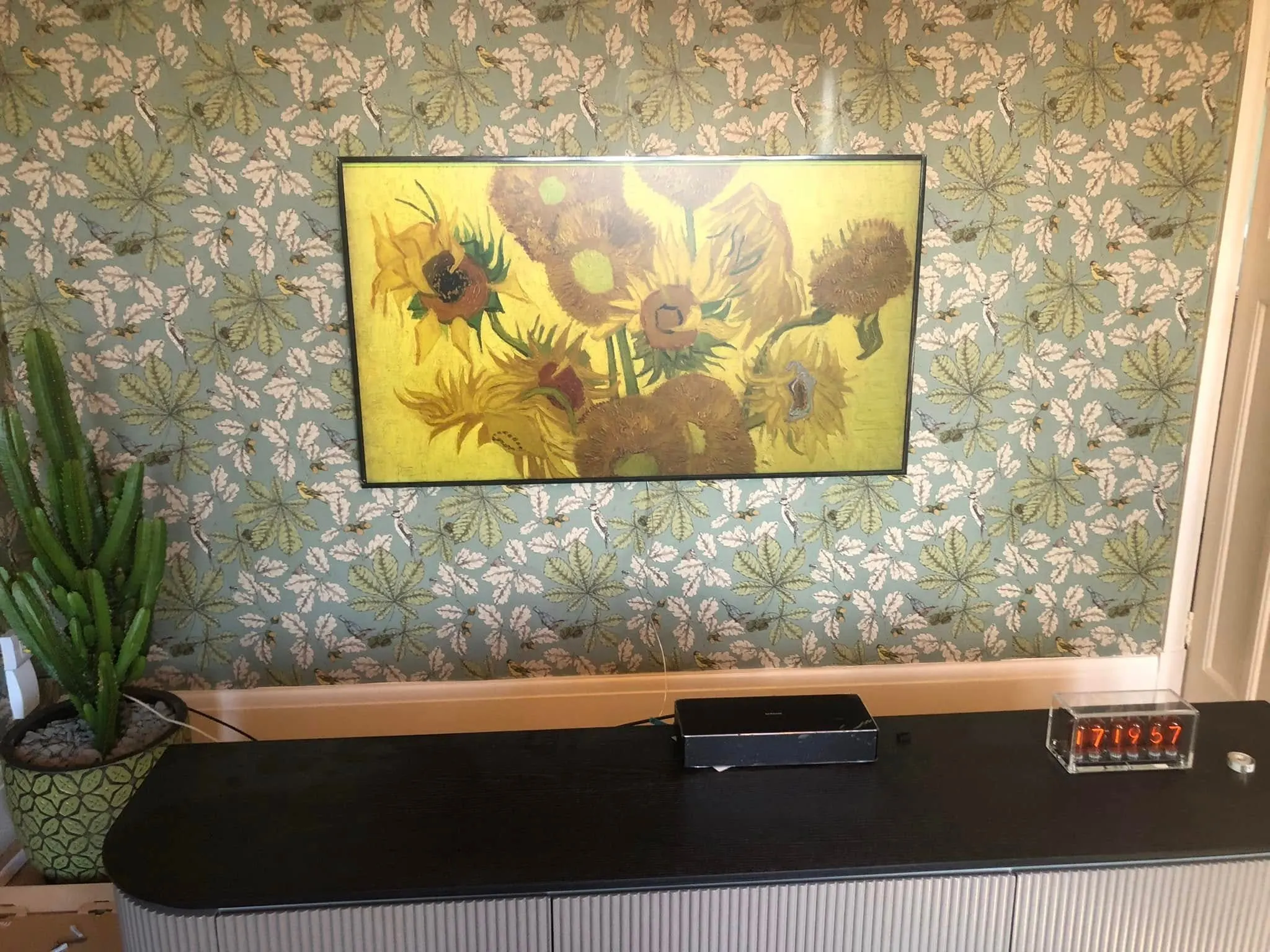 Wall-mounted TV displaying a sunflower painting on leaf-patterned wallpaper above a black-topped sideboard with a nixie clock
